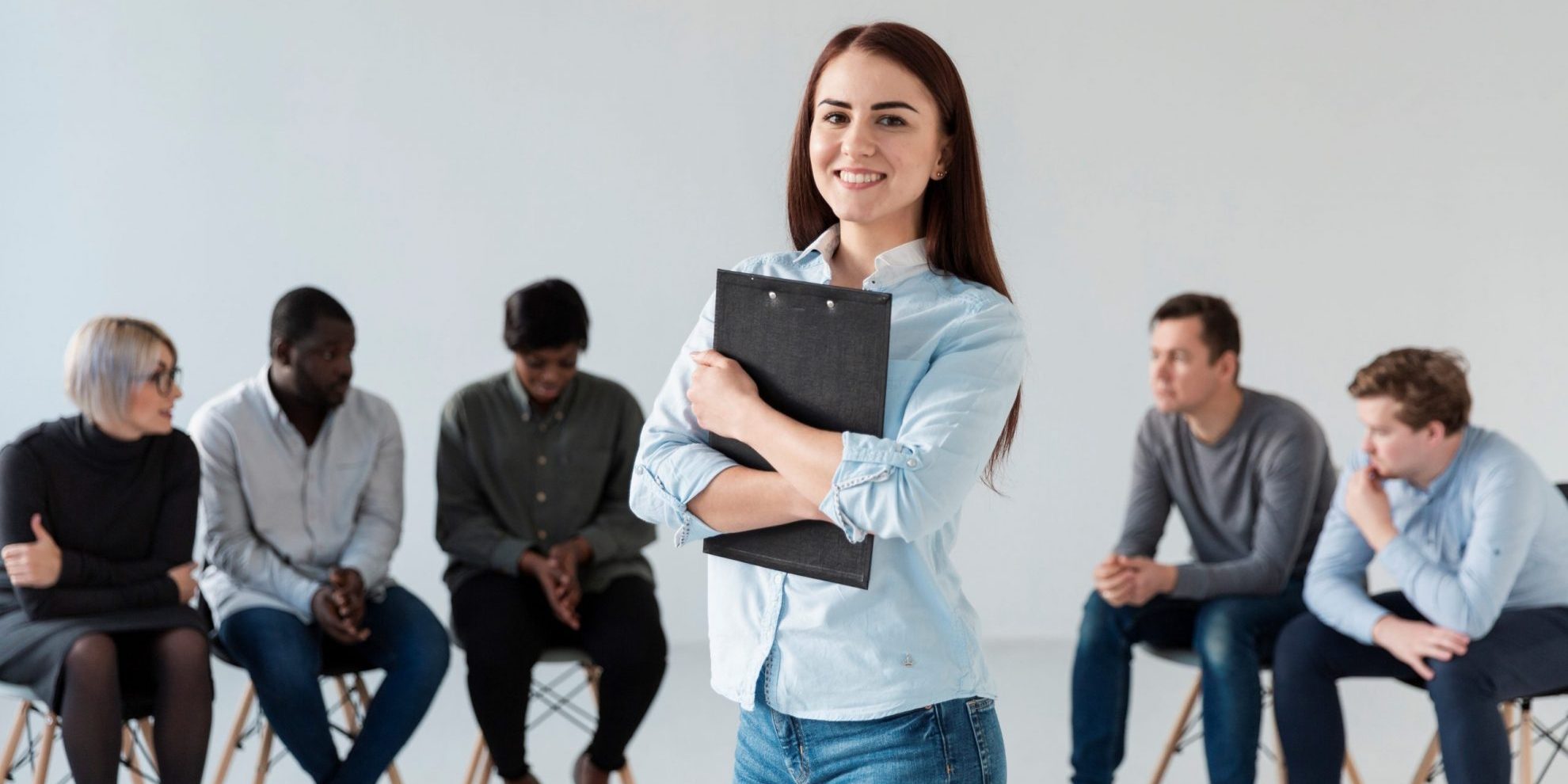Smiling woman holding clipboard with people sitting in the background illustrates blog "Active vs Passive Candidates: What Is the Difference?"