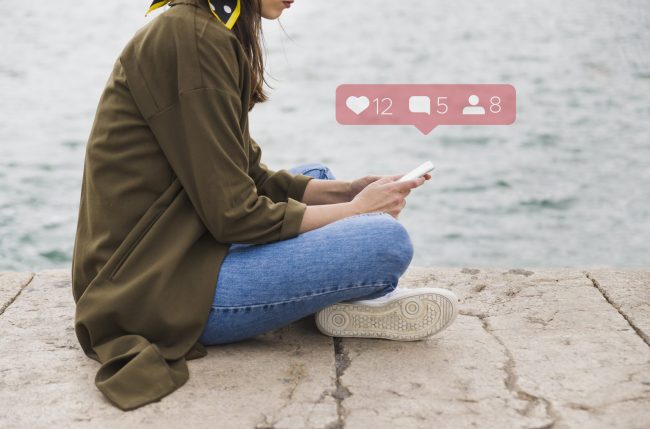 social-media-fp Woman wearing jeans and a green jacket sitting on the floor holding smartphone with social media icons hovering above it.